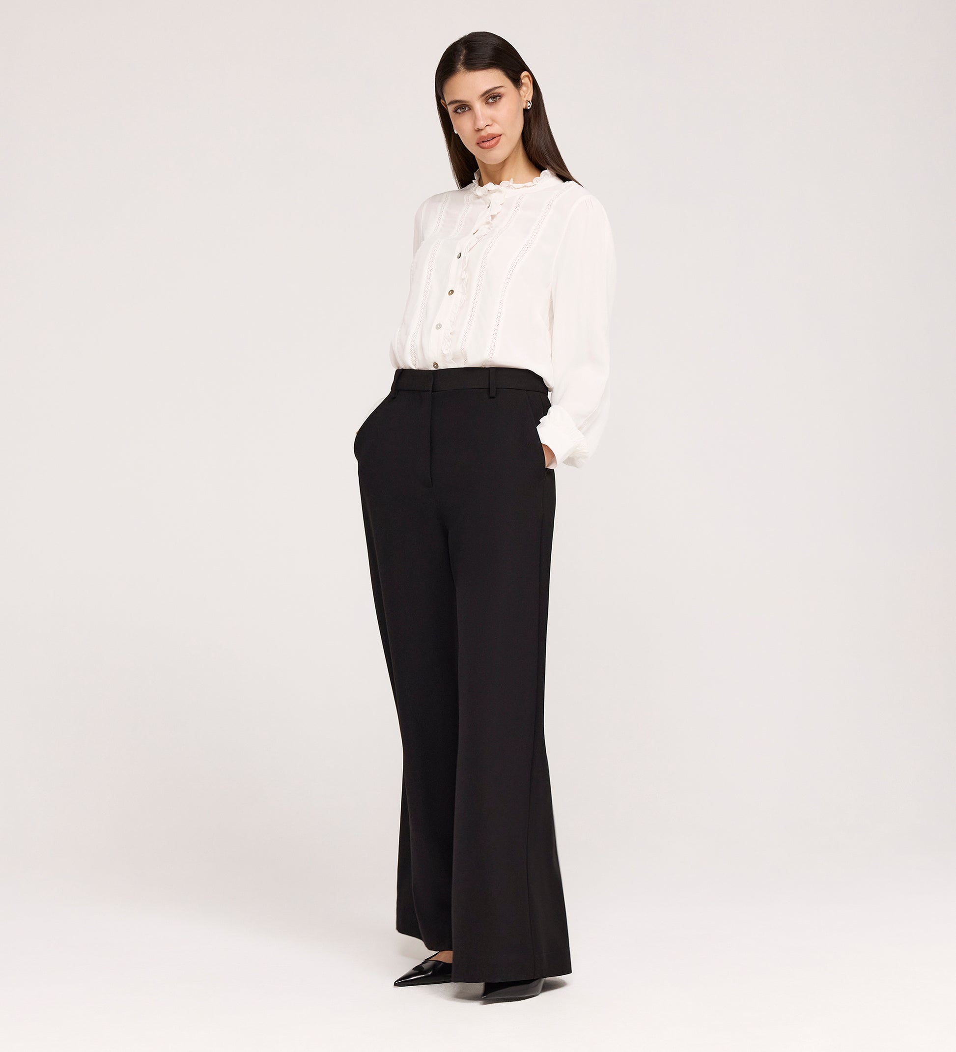 A woman stands against a plain background, wearing high-waisted Addie Black Gaberdine Twill Wide Leg Trousers, a white ruffle long-sleeve blouse, and black pointed-toe shoes with her hands in her pockets.