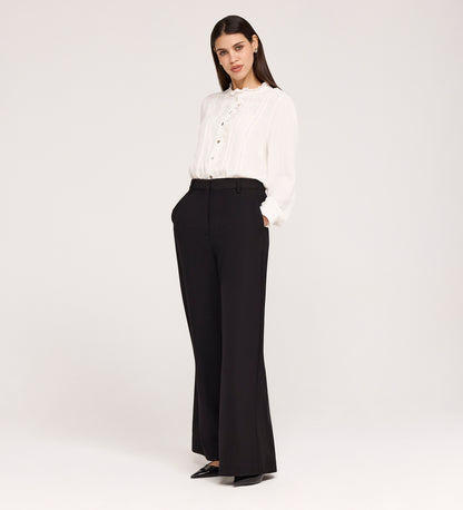 A woman stands against a plain background, wearing high-waisted Addie Black Gaberdine Twill Wide Leg Trousers, a white ruffle long-sleeve blouse, and black pointed-toe shoes with her hands in her pockets.