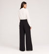 A woman faces away, wearing a long-sleeve white blouse and Addie Black Gaberdine Twill Wide Leg Trousers with black shoes, set against a plain light background.