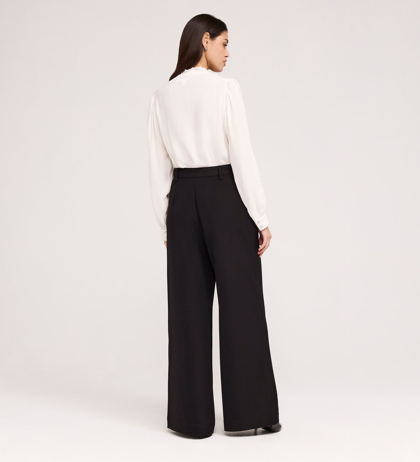 A woman faces away, wearing a long-sleeve white blouse and Addie Black Gaberdine Twill Wide Leg Trousers with black shoes, set against a plain light background.