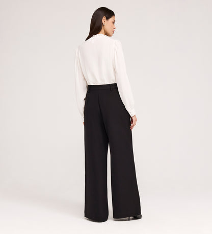 A woman faces away, wearing a long-sleeve white blouse and Addie Black Gaberdine Twill Wide Leg Trousers with black shoes, set against a plain light background.