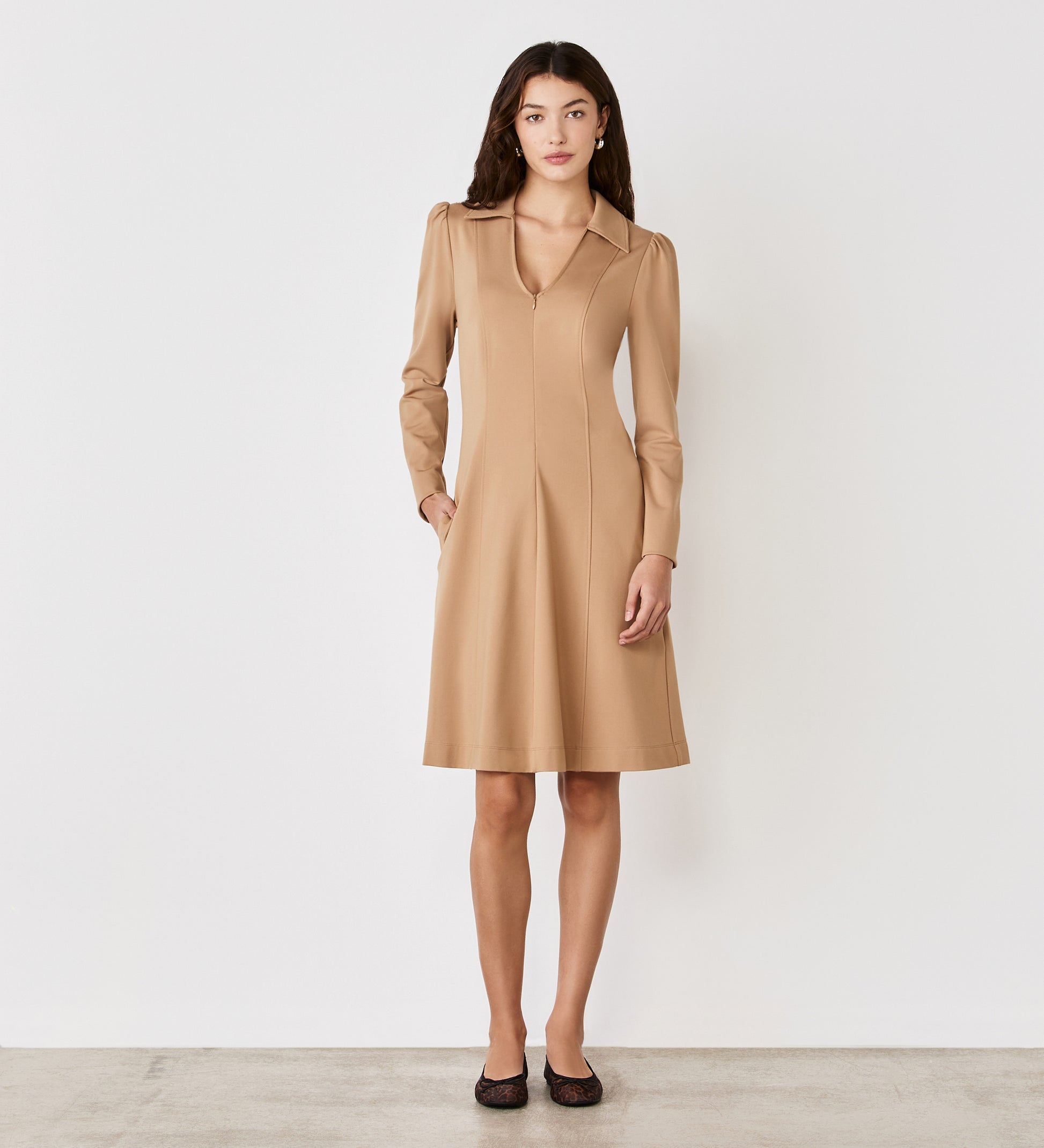 A woman with long dark hair and a neutral expression stands against a plain white wall, wearing the Bethany Camel Ponte Jersey Knee Length Dress with long sleeves, a collar, and black flats.