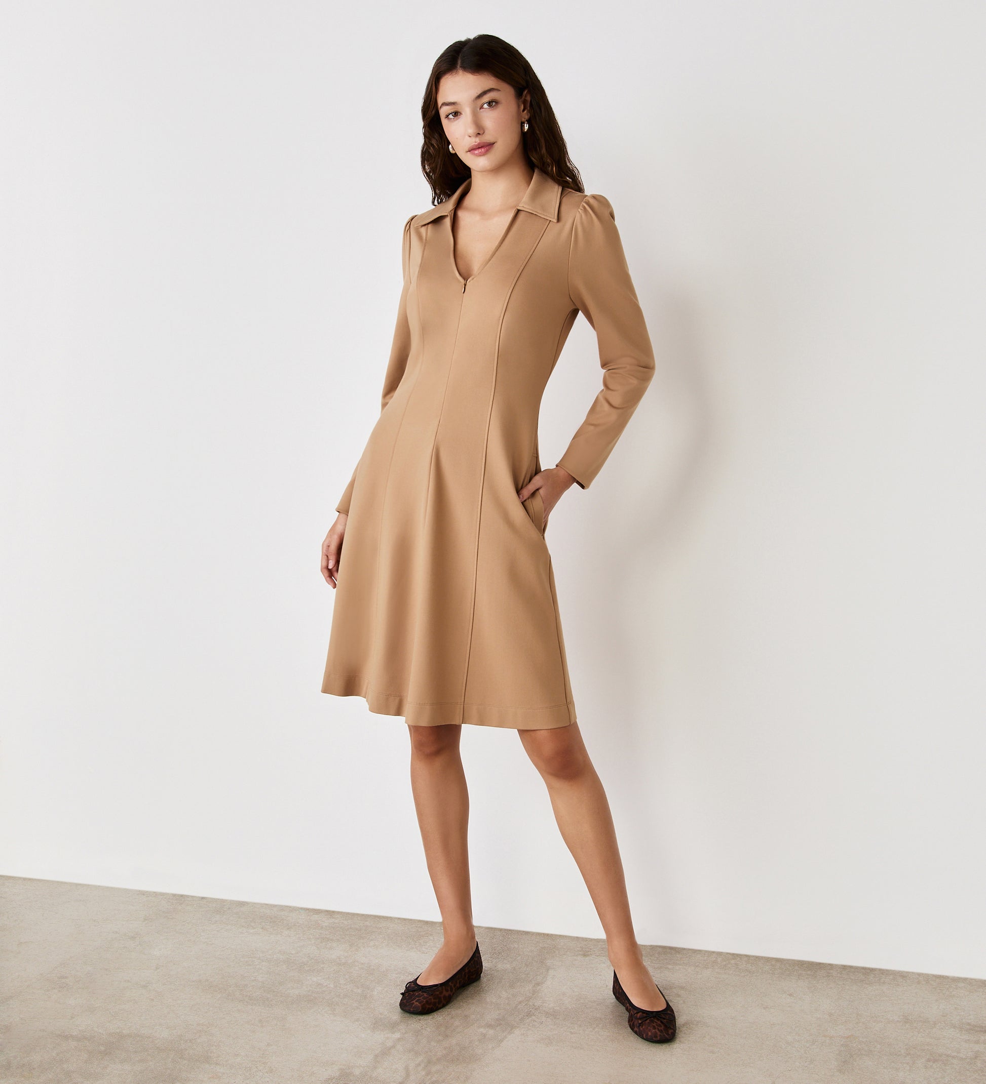 A woman poses confidently against a white wall in the Bethany Camel Ponte Jersey Knee Length Dress, featuring a collar, long sleeves, pockets, and black flats. The soft jersey fabric adds comfort to its relaxed elegance.