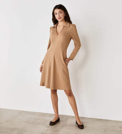 A woman poses confidently against a white wall in the Bethany Camel Ponte Jersey Knee Length Dress, featuring a collar, long sleeves, pockets, and black flats. The soft jersey fabric adds comfort to its relaxed elegance.