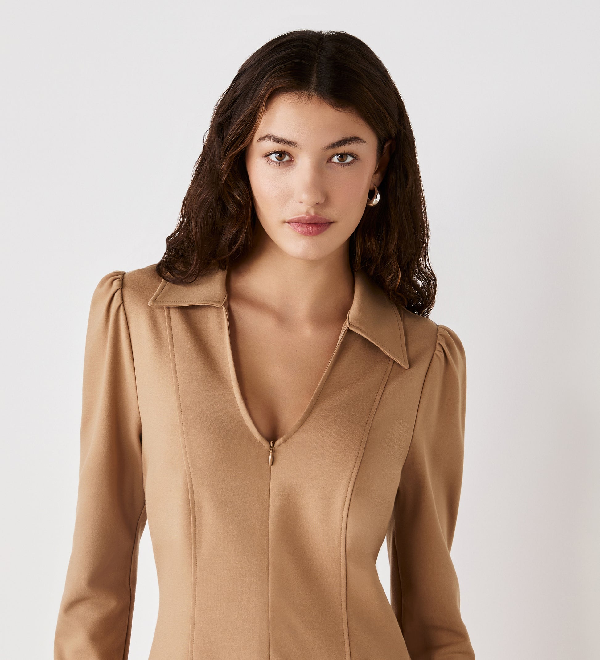 A woman with wavy dark hair, wearing the Bethany Camel Ponte Jersey Knee Length Dress with long sleeves and a subtle v-neck, stands against a plain white background and looks directly at the camera.