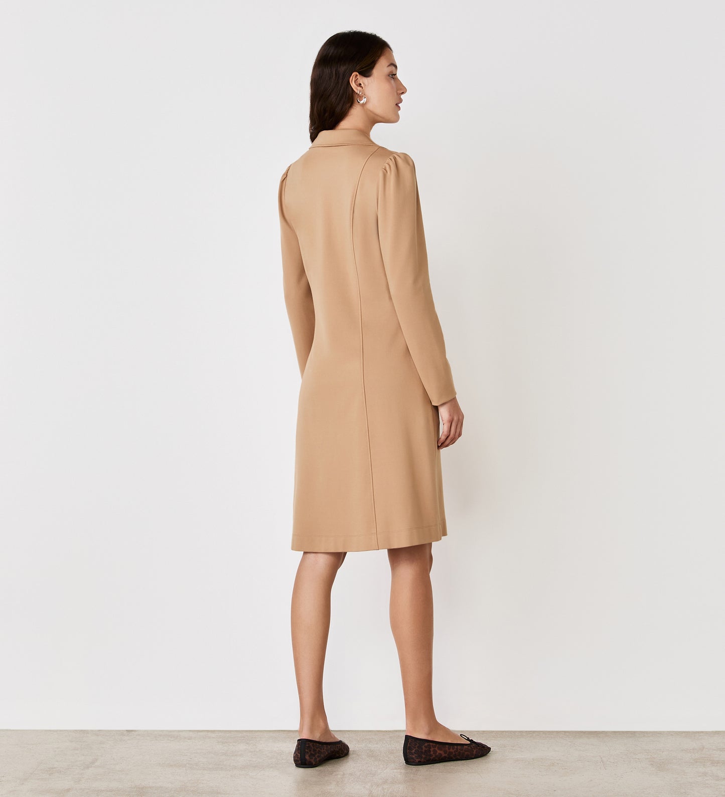 A woman stands with her back to the camera, wearing the Bethany Camel Ponte Jersey Knee Length Dress and black flats, set against a plain white background.