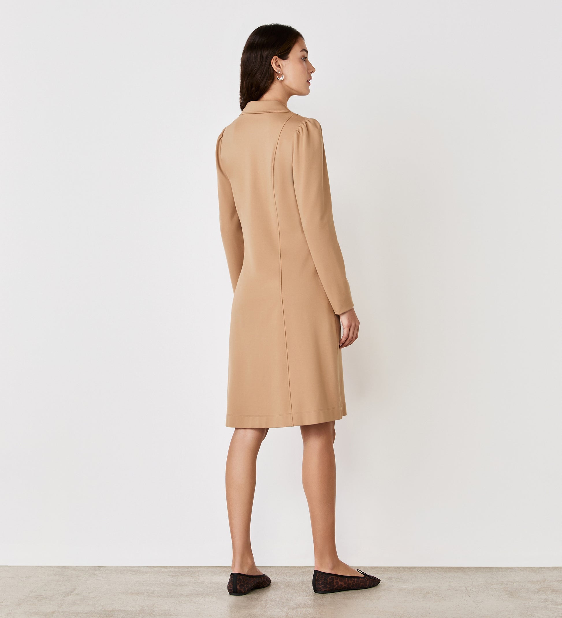 A woman stands with her back to the camera, wearing the Bethany Camel Ponte Jersey Knee Length Dress and black flats, set against a plain white background.