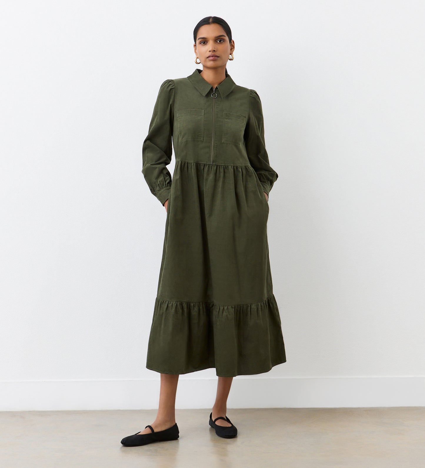 A woman stands in front of a white wall, confidently looking at the camera with her hands in the pockets of the Callie Khaki Corduroy Midi Dress, featuring long sleeves, a collared neckline, a tiered hemline, and paired with black flats.