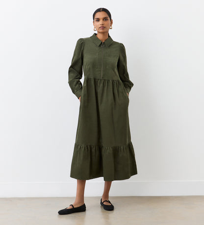 A woman stands in front of a white wall, confidently looking at the camera with her hands in the pockets of the Callie Khaki Corduroy Midi Dress, featuring long sleeves, a collared neckline, a tiered hemline, and paired with black flats.
