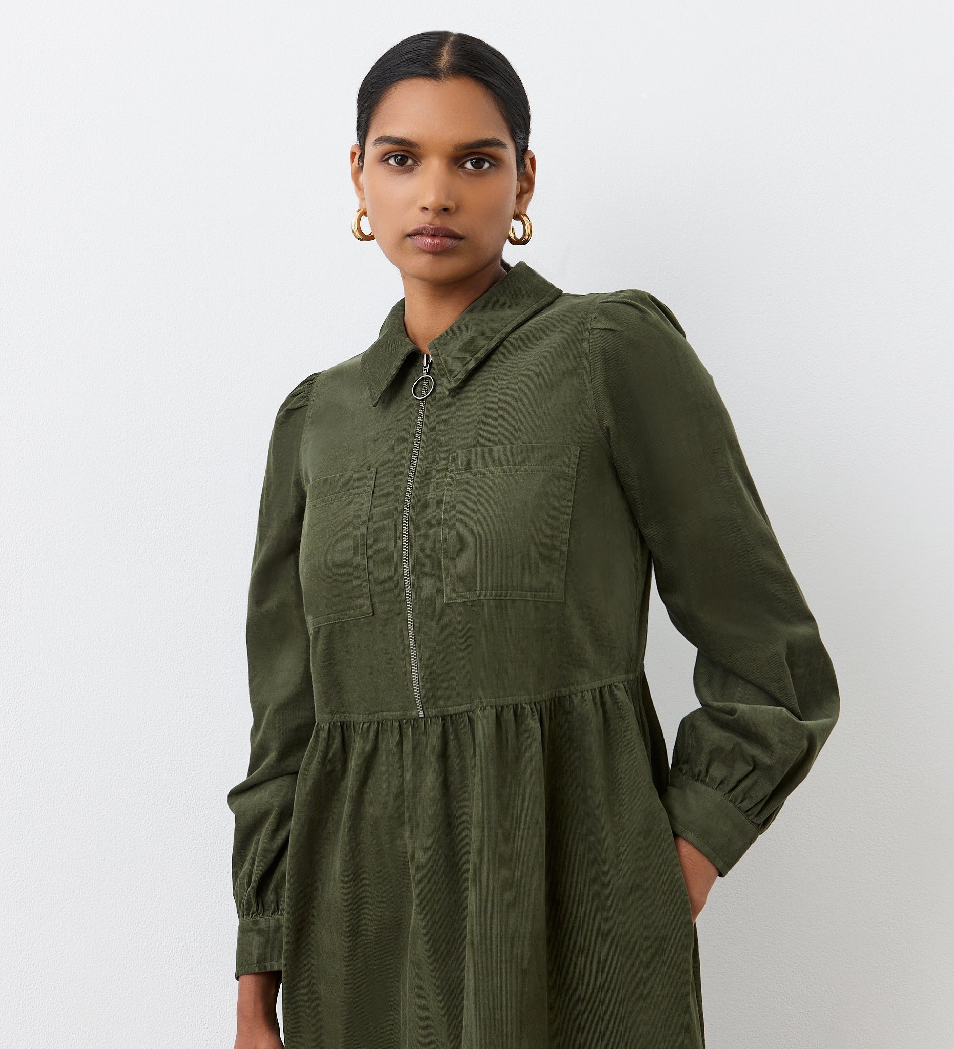A woman confidently stands against a plain white background wearing the Callie Khaki Corduroy Midi Dress, featuring chest pockets and a front zipper.
