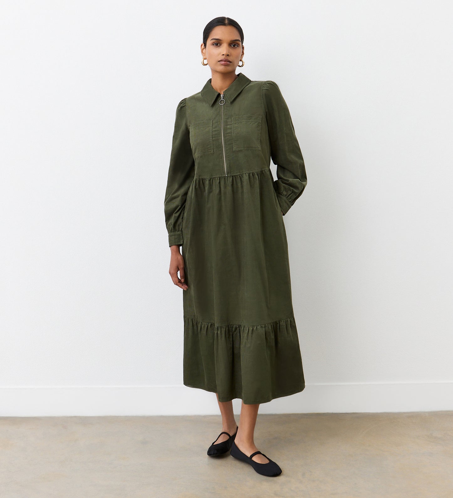 A woman with her hair tied back stands against a plain white wall, wearing the Callie Khaki Corduroy Midi Dress with long sleeves and a collar, paired with black flats. She faces the camera with a neutral expression.