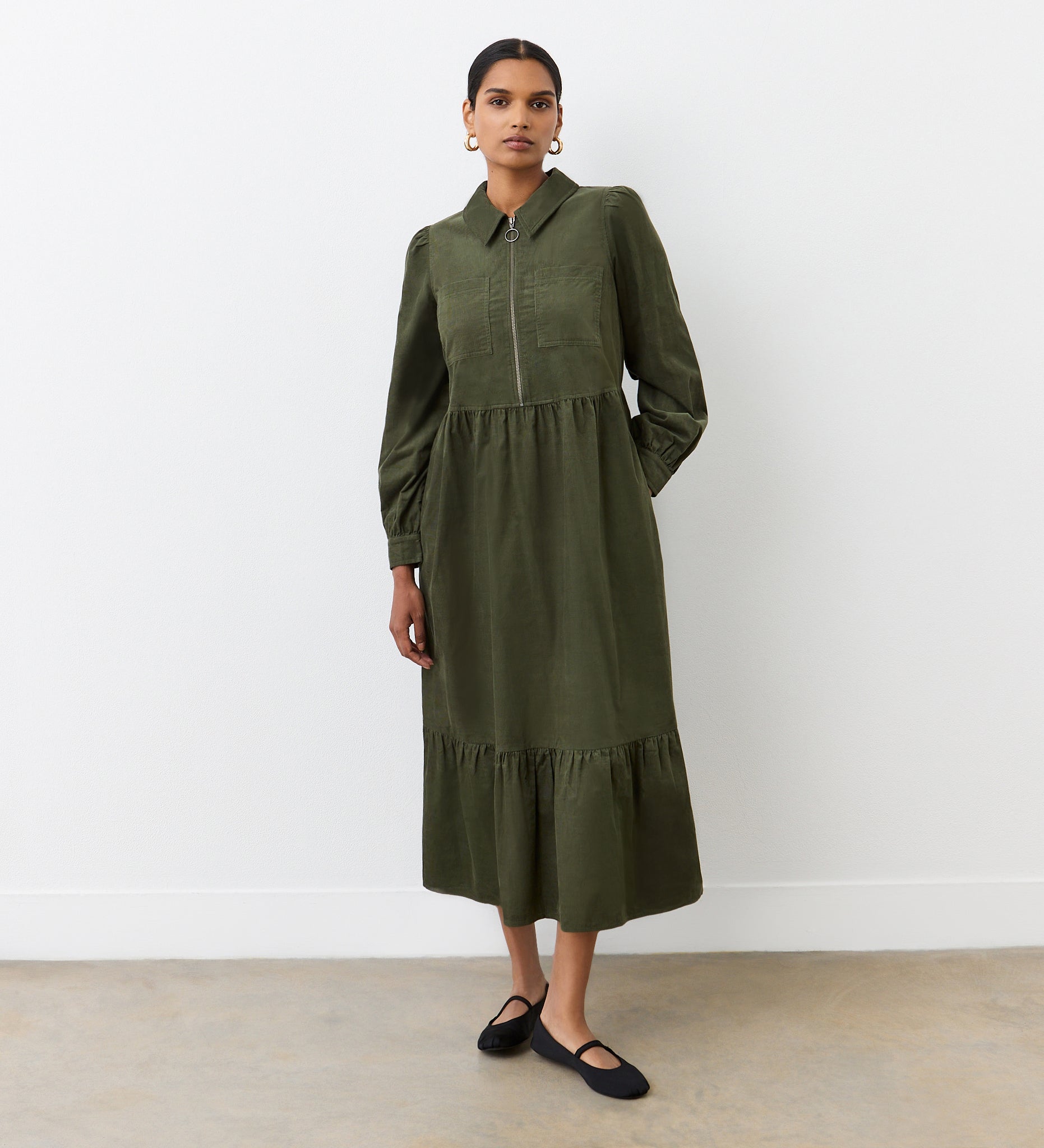 A woman with her hair tied back stands against a plain white wall, wearing the Callie Khaki Corduroy Midi Dress with long sleeves and a collar, paired with black flats. She faces the camera with a neutral expression.