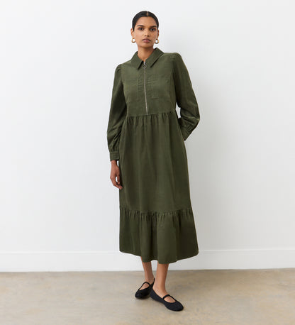 A woman with her hair tied back stands against a plain white wall, wearing the Callie Khaki Corduroy Midi Dress with long sleeves and a collar, paired with black flats. She faces the camera with a neutral expression.