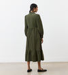 A woman with dark hair tied back, wearing the Callie Khaki Corduroy Midi Dress with long sleeves and a tiered hemline, stands indoors in front of a white wall facing away. She pairs the dress with black flat shoes.