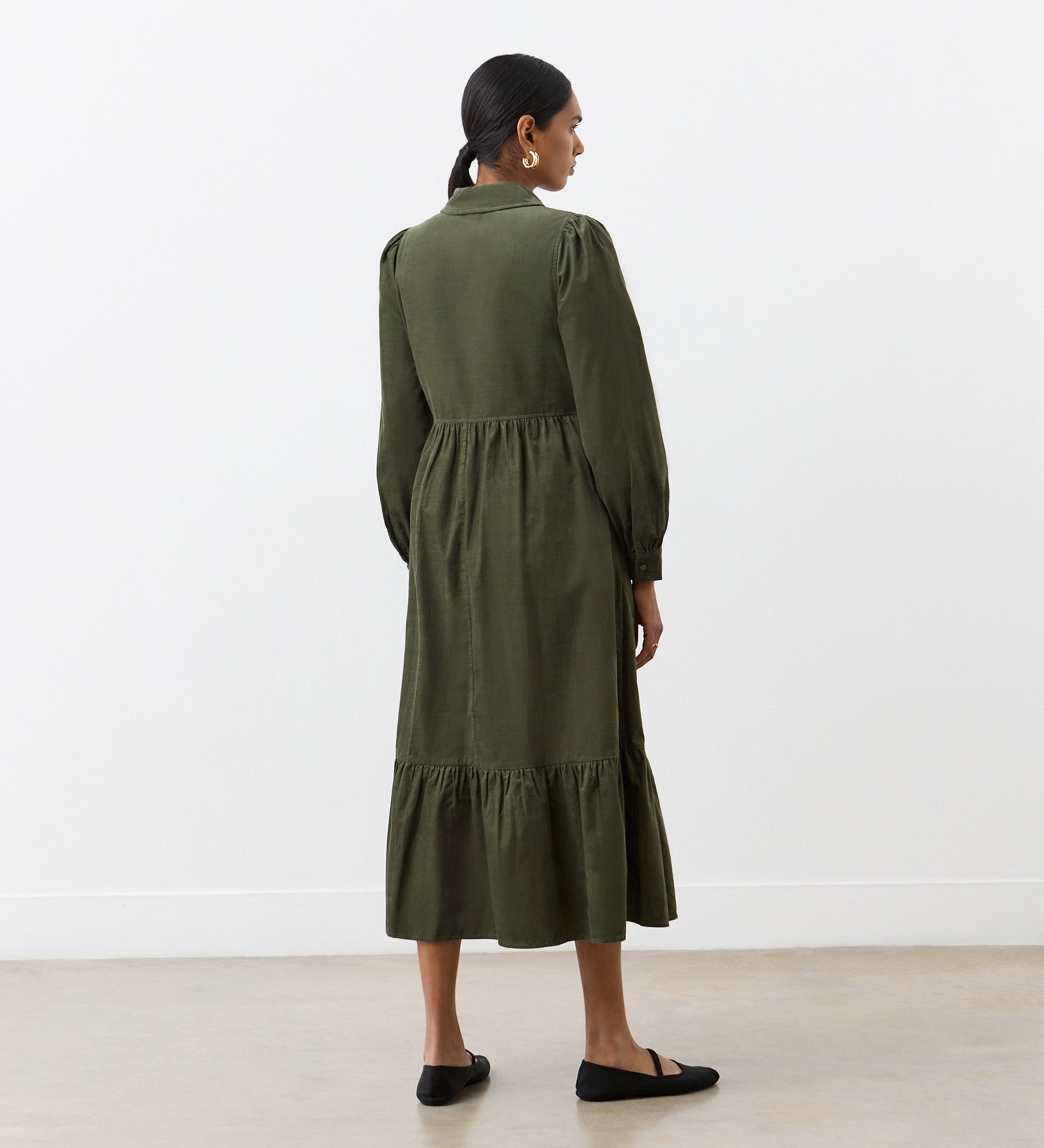 A woman with dark hair tied back, wearing the Callie Khaki Corduroy Midi Dress with long sleeves and a tiered hemline, stands indoors in front of a white wall facing away. She pairs the dress with black flat shoes.