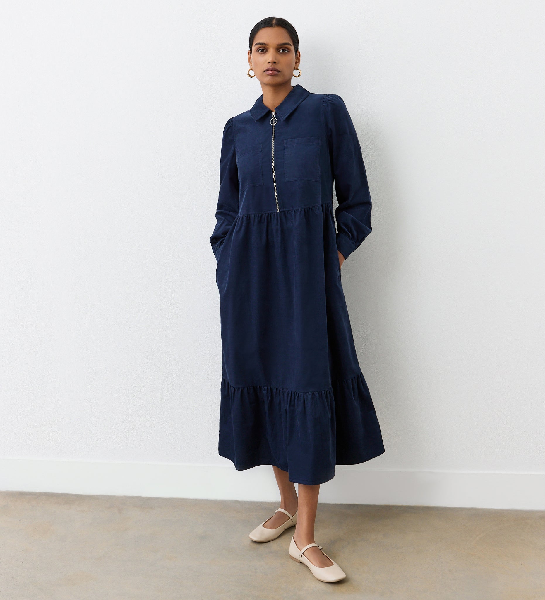 A woman stands against a white wall wearing the Callie Navy Corduroy Midi Dress, featuring a collared neckline, tiered hemline, and pockets. She pairs it with beige flats and hoop earrings.