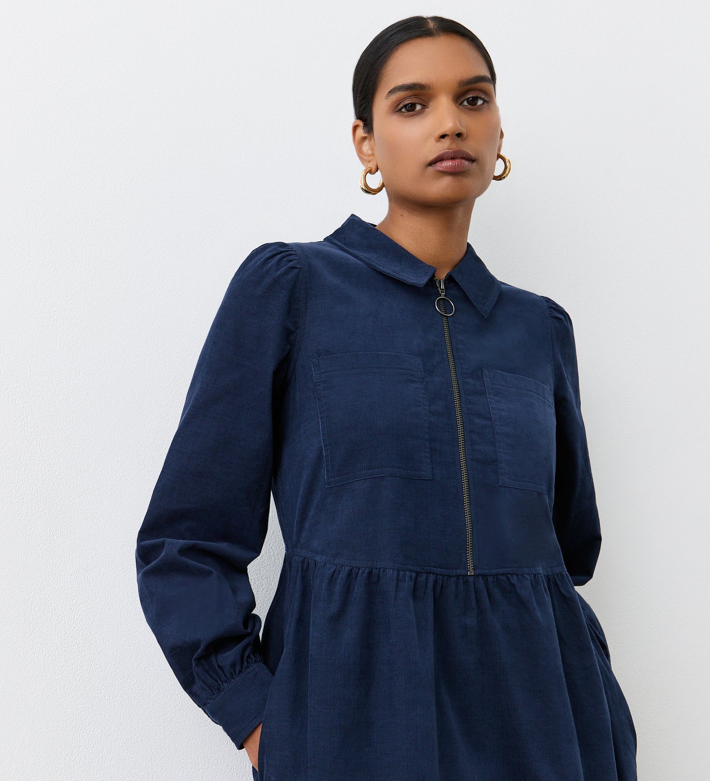 A woman with dark hair pulled back models the Callie Navy Corduroy Midi Dress, featuring a collared neckline, zip front, and chest pockets. She stands against a white background, looking confidently at the camera.