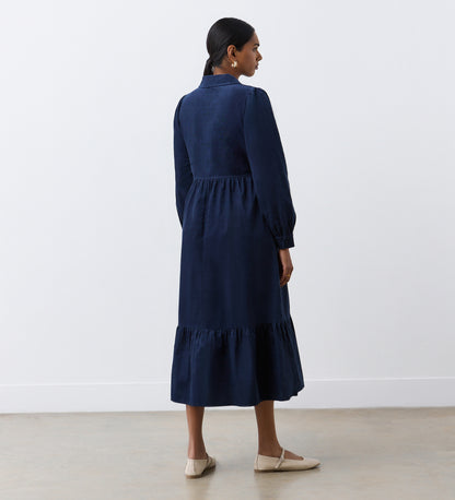 A woman with dark hair tied back wears the Callie Navy Corduroy Midi Dress, featuring a gathered waist and tiered hemline, paired with light-colored flats. She stands indoors facing away against a plain white background.