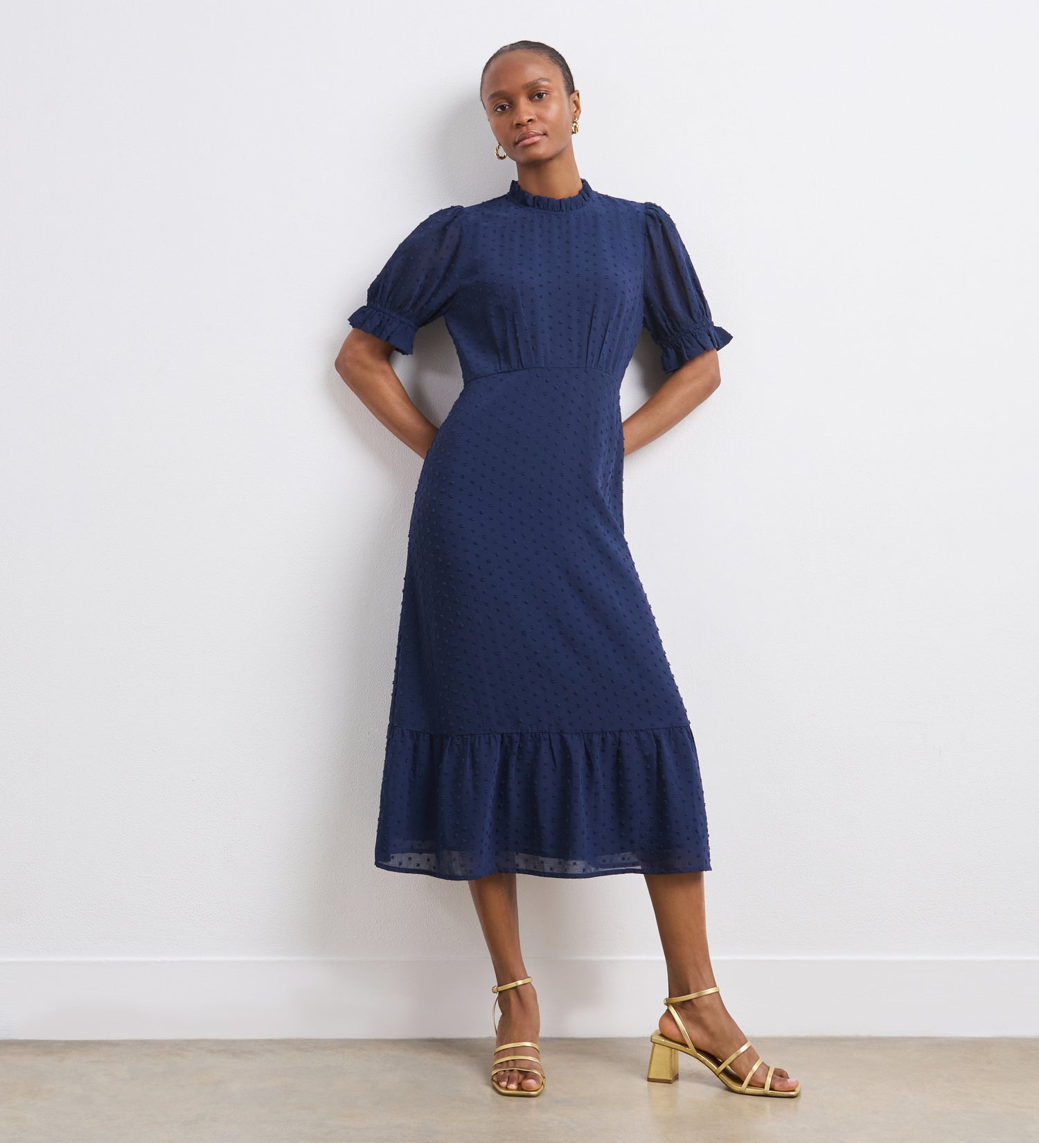A woman with short hair wears the Camille Navy Chiffon Midi Dress, featuring short puff sleeves, a ruffle neck, and tiered hem. She pairs it with gold strappy sandals and hoop earrings, standing against a white wall.
