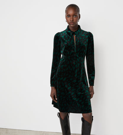 A woman stands against a white wall wearing the Capri Green Animal Knee Length Dress featuring a collar, keyhole neckline, and empire waistband, paired with black knee-high boots—perfect for party season.
