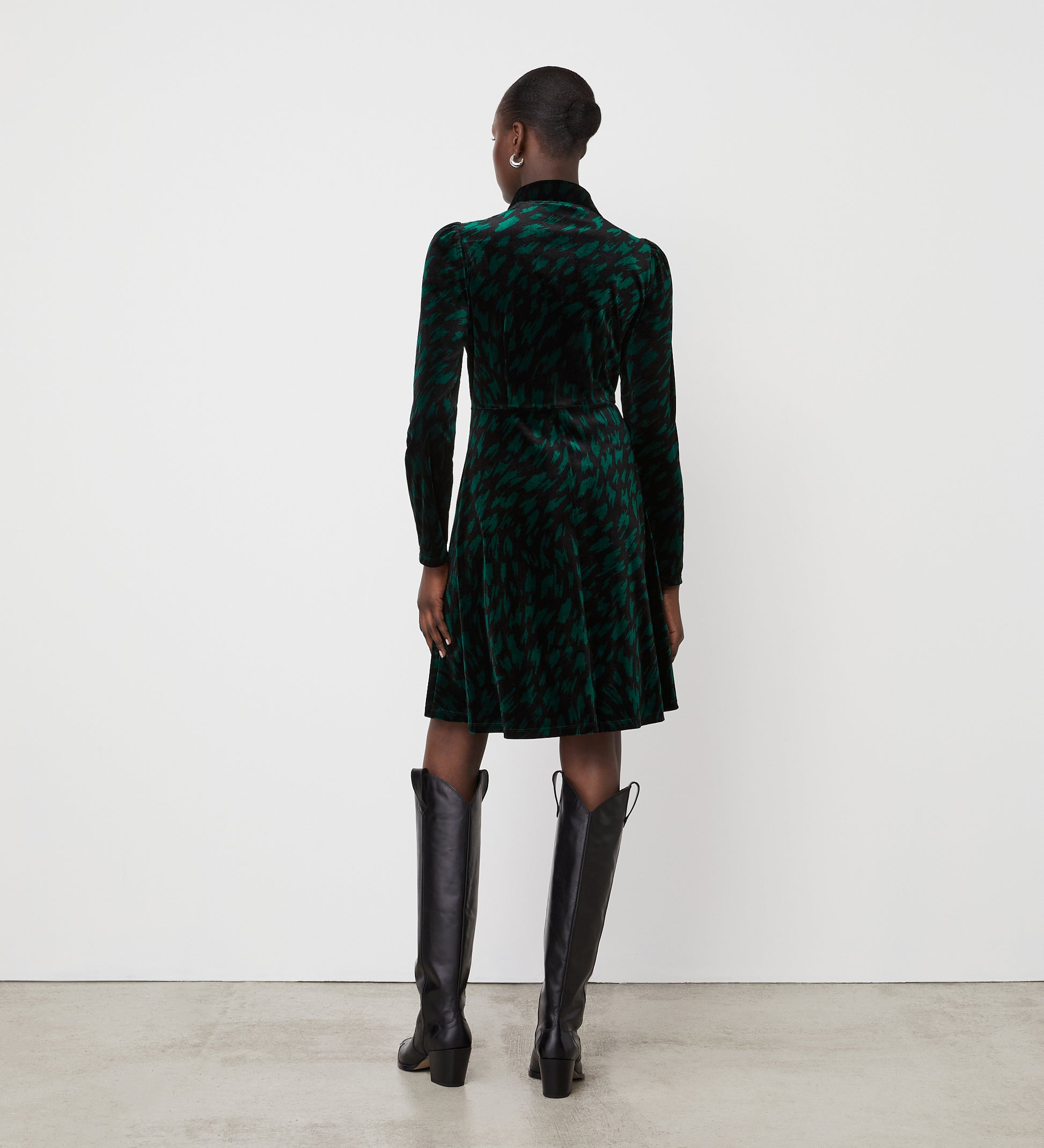 A woman faces away from the camera in a Capri Green Animal Knee Length Dress with an empire waistband and tall black boots. Her hair is styled in a low bun as she stands against a plain white wall and concrete floor.