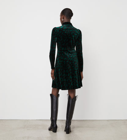 A woman faces away from the camera in a Capri Green Animal Knee Length Dress with an empire waistband and tall black boots. Her hair is styled in a low bun as she stands against a plain white wall and concrete floor.