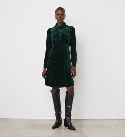 A woman stands against a plain white wall wearing the Capri Green Animal Knee Length Dress with long sleeves, a collared neckline, and an empire waistband, paired with black knee-high boots. She faces forward with her arms relaxed at her sides.