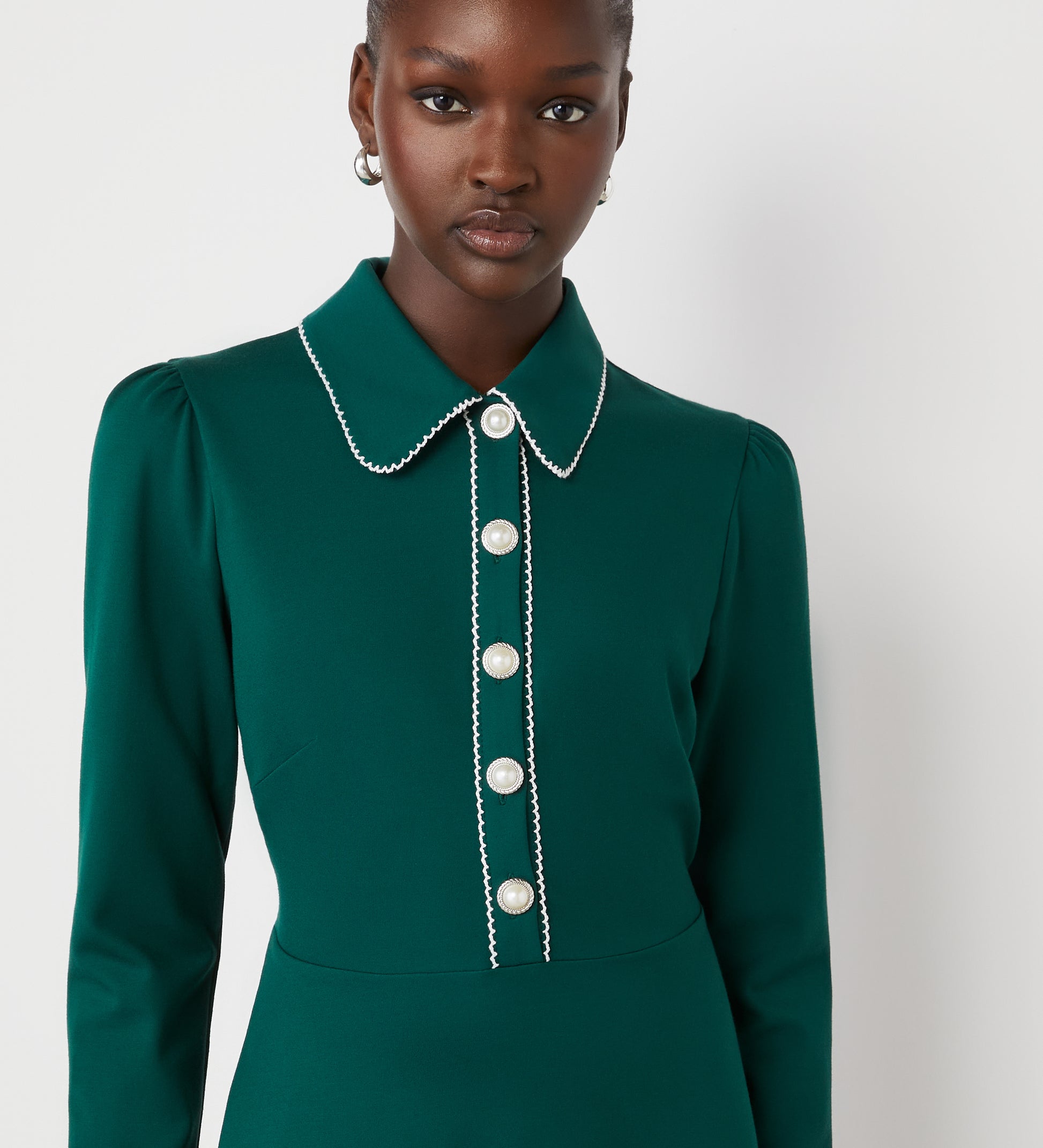 A woman stands against a plain light background wearing the Clarice Bottle Green Ponte Jersey Midi Dress, featuring long sleeves, pearl-like buttons on the functional placket, and white trim along the collar and placket.
