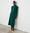 Wearing the Clarice Bottle Green Ponte Jersey Midi Dress with long sleeves, a white collar, and functional placket, a person stands against a white wall, styled with light-colored pointed heels.