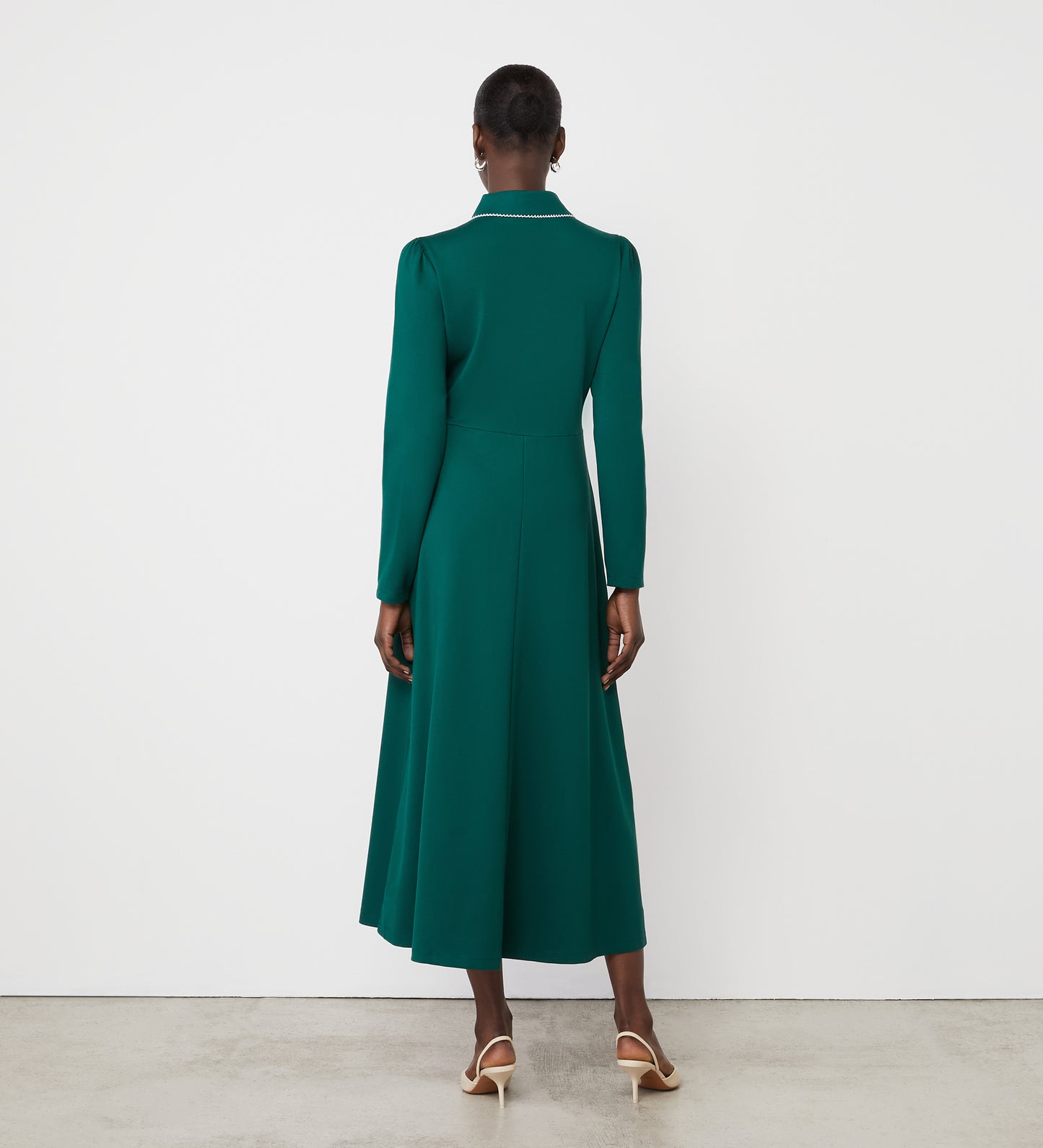 A woman with short hair stands with her back to the camera on a bare floor, wearing the Clarice Bottle Green Ponte Jersey Midi Dress with long sleeves and a functional placket, paired with white heeled shoes against a white wall.