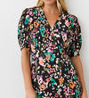 A woman models the Colette Leopard Crepe Midi Tea Dress—a black dress with vibrant floral and animal motifs, short puff sleeves, V-neckline, and empire waist—made from crease-free fabric, standing before a plain white background.