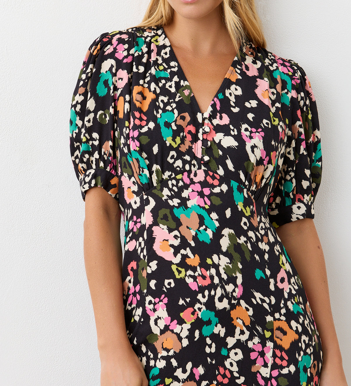 A woman models the Colette Leopard Crepe Midi Tea Dress—a black dress with vibrant floral and animal motifs, short puff sleeves, V-neckline, and empire waist—made from crease-free fabric, standing before a plain white background.