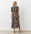 A woman faces away, wearing the Colette Leopard Crepe Midi Tea Dress with an empire waist. She has blonde hair, white strappy high-heeled sandals, and stands in a plain indoor setting.