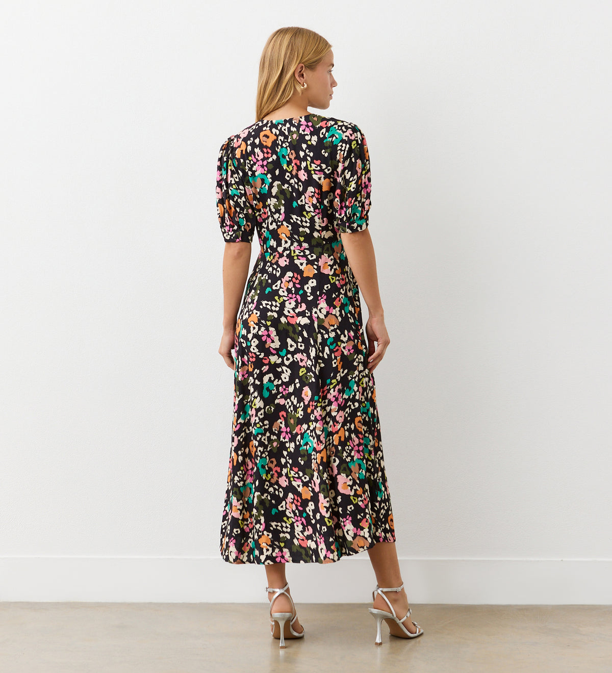 A woman faces away, wearing the Colette Leopard Crepe Midi Tea Dress with an empire waist. She has blonde hair, white strappy high-heeled sandals, and stands in a plain indoor setting.