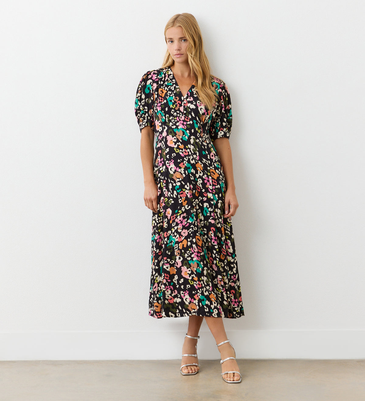 A woman with long blonde hair and a relaxed expression stands against a white wall wearing the Colette Leopard Crepe Midi Tea Dress and silver strappy sandals.