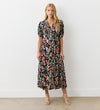 A woman stands by a white wall wearing the Colette Leopard Crepe Midi Tea Dress with short puff sleeves and strappy silver heels. Her long blonde hair is loose.