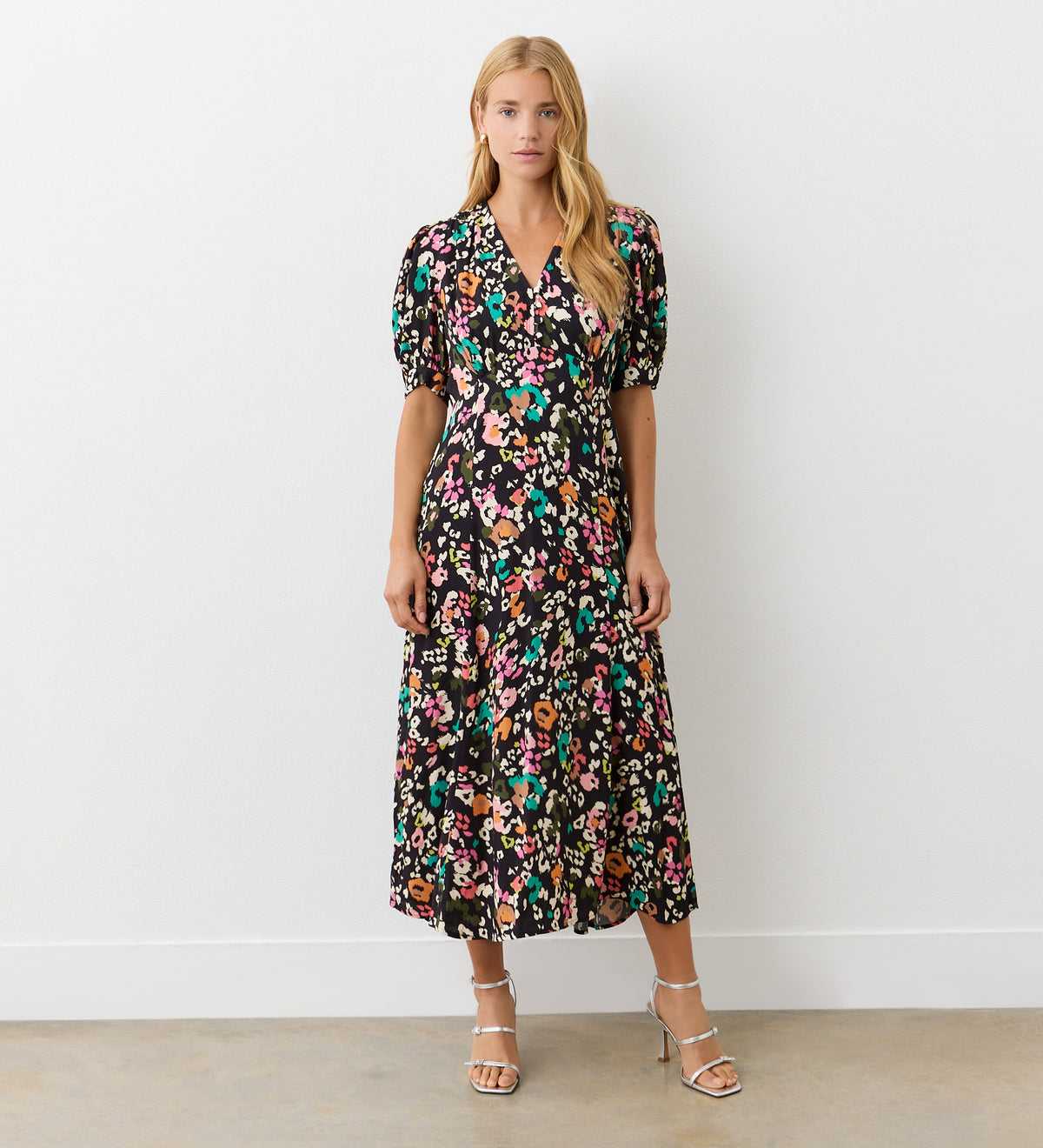 A woman stands by a white wall wearing the Colette Leopard Crepe Midi Tea Dress with short puff sleeves and strappy silver heels. Her long blonde hair is loose.