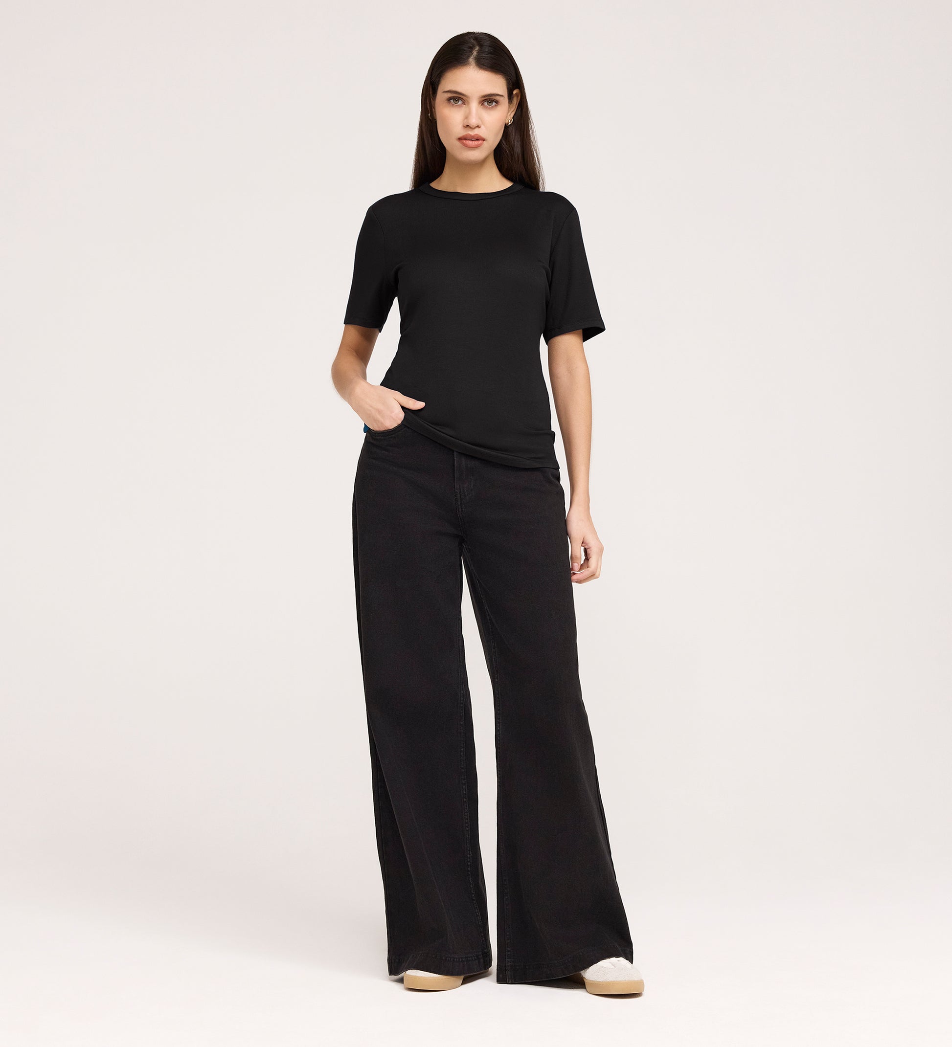 A woman stands against a plain background wearing the Cherry Black Short Sleeve Jersey T-Shirt and wide-leg black jeans, her left hand in her pocket as she looks directly at the camera.