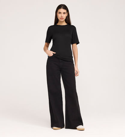 A woman stands against a plain background wearing the Cherry Black Short Sleeve Jersey T-Shirt and wide-leg black jeans, her left hand in her pocket as she looks directly at the camera.