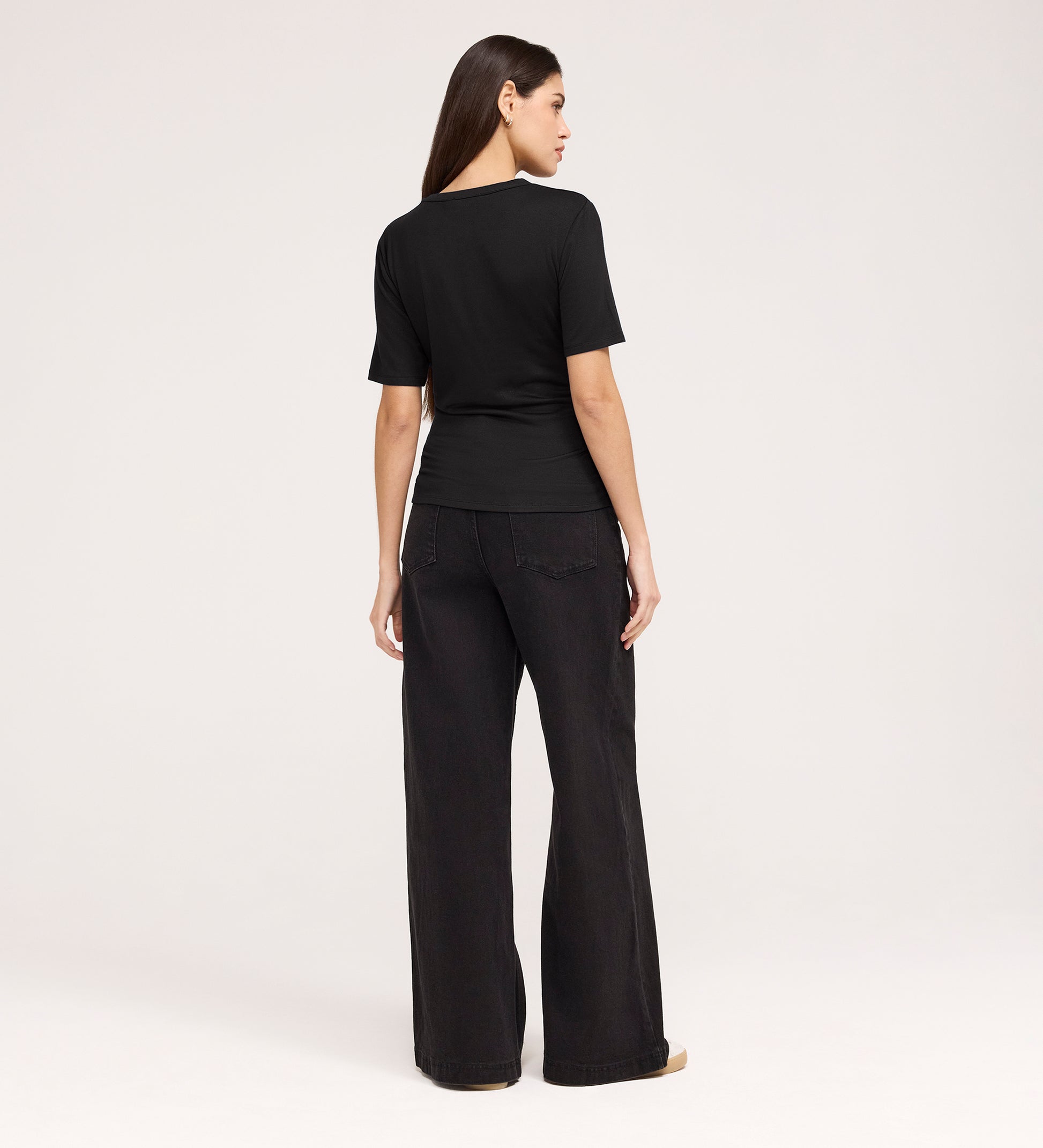 A woman with long brown hair, facing away against a light background, wears the Cherry Black Short Sleeve Jersey T-Shirt and wide-leg black jeans.