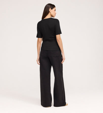 A woman with long brown hair, facing away against a light background, wears the Cherry Black Short Sleeve Jersey T-Shirt and wide-leg black jeans.