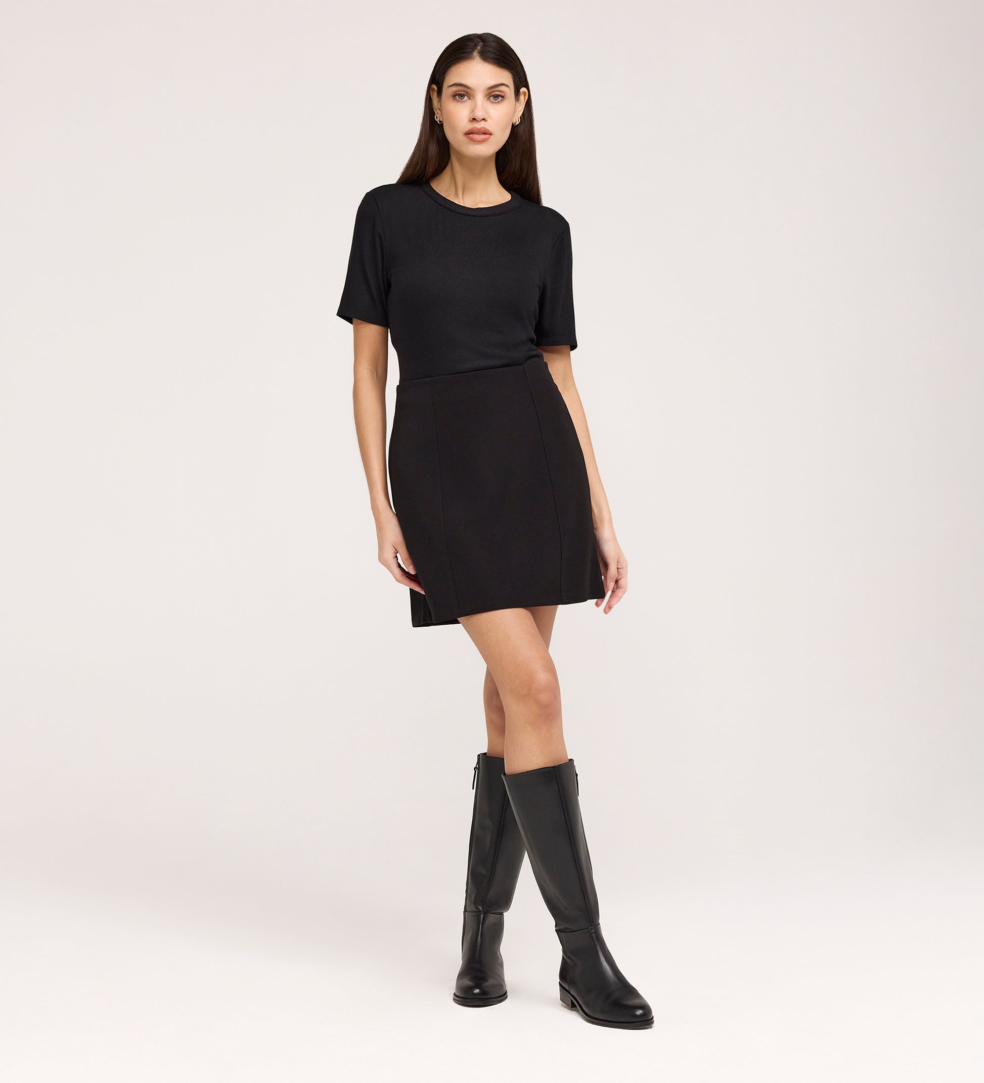 A woman stands against a plain background, wearing the Cherry Black Short Sleeve Jersey T-Shirt with a relaxed fit, paired with a black mini skirt and knee-high black boots.
