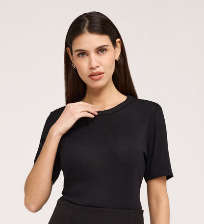 A woman with long brown hair stands against a light background, gently touching her collar while wearing the Cherry Black Short Sleeve Jersey T-Shirt. The plain black top is made from rib jersey fabric, and she gazes forward with a neutral expression.