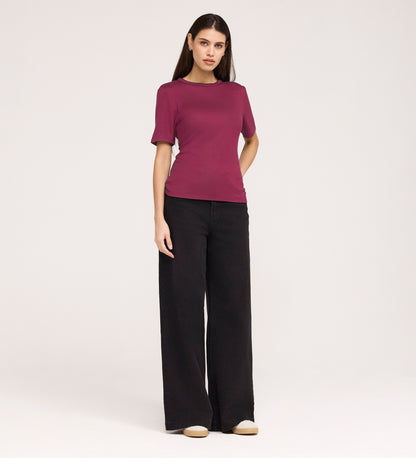 A woman with long straight hair and a neutral expression stands against a plain background, wearing the Cherry Pink Short Sleeve Jersey T-Shirt, wide-leg black pants, and beige shoes.