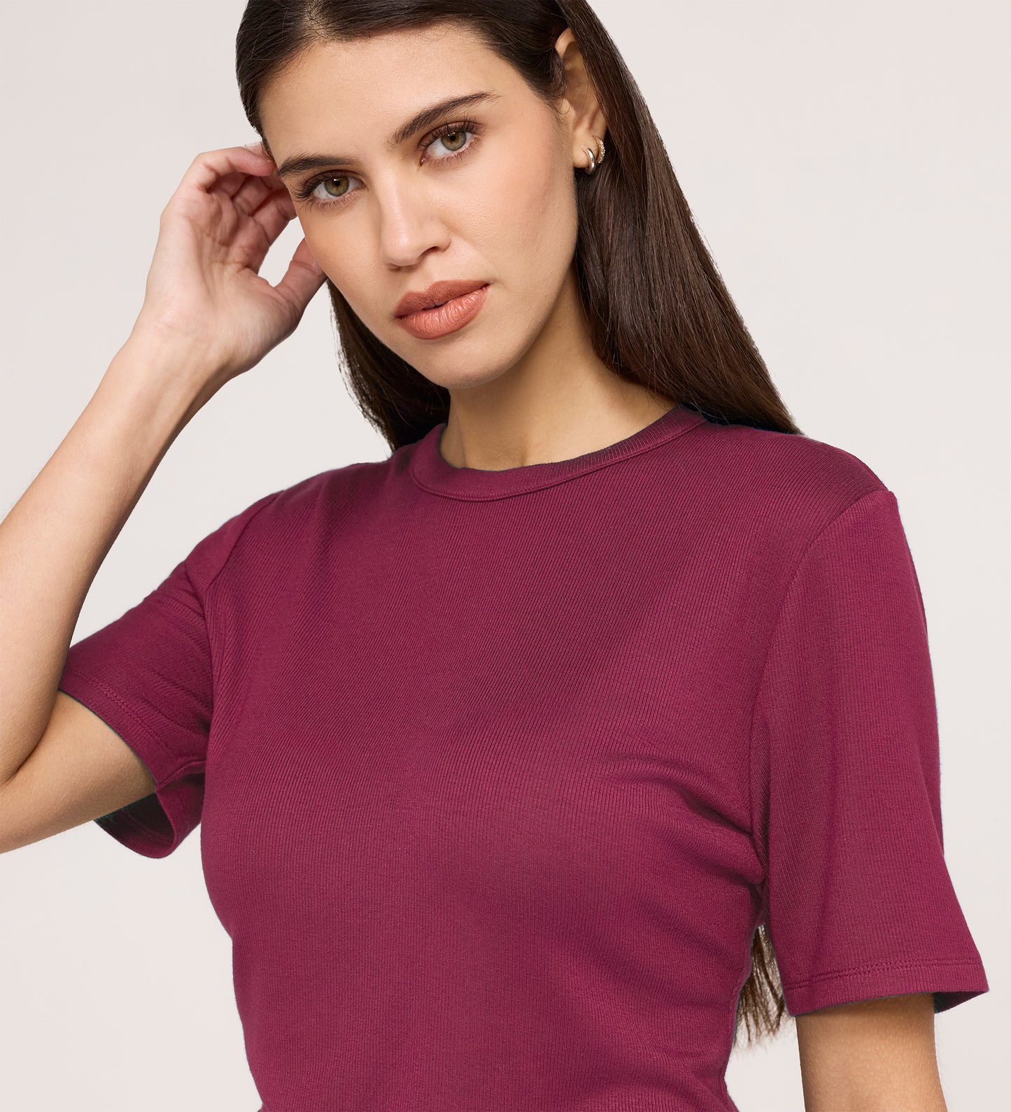 A woman with long brown hair wears a Cherry Pink Short Sleeve Jersey T-Shirt, standing against a light background, touching her hair with one hand and looking at the camera with a neutral expression.
