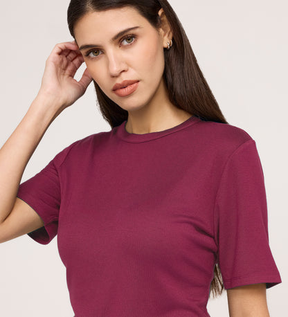 A woman with long brown hair wears a Cherry Pink Short Sleeve Jersey T-Shirt, standing against a light background, touching her hair with one hand and looking at the camera with a neutral expression.