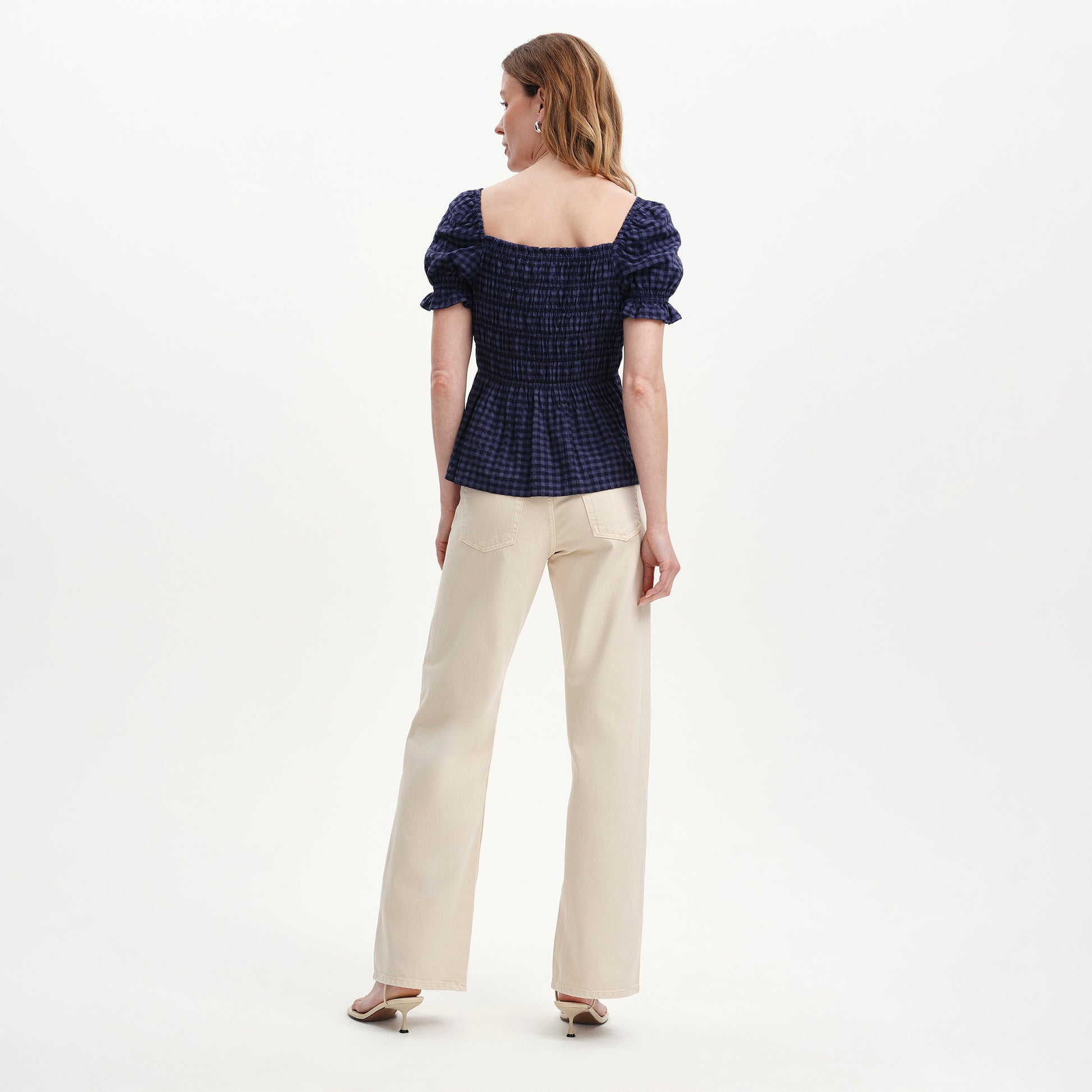 Woman wearing Cynthia Navy Gingham Seersucker Shirred Puff Sleeve Top and beige pants on a white background