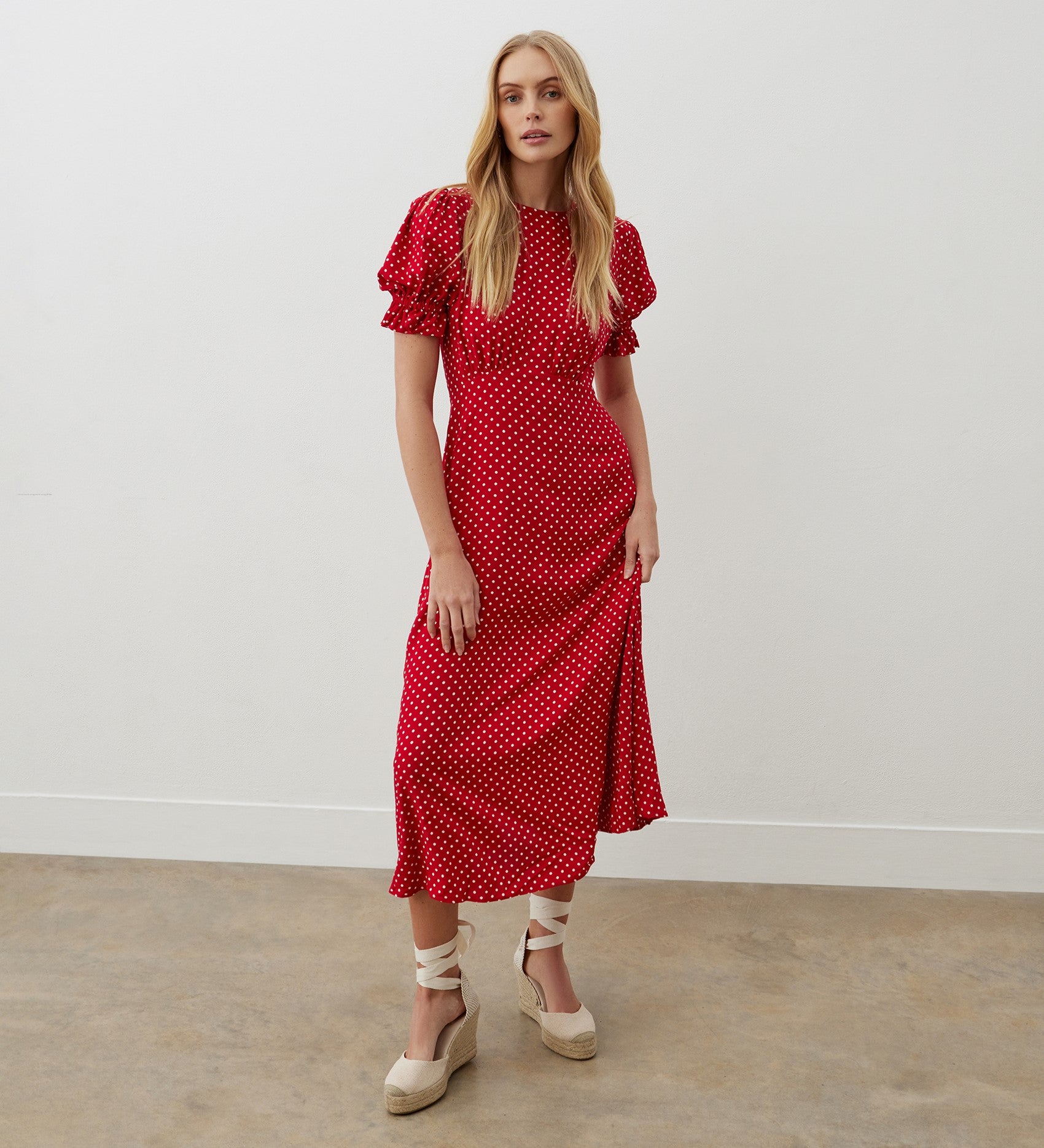 A woman stands indoors facing forward against a white wall and concrete floor, wearing the Mela Red Polka Dot Crepe Midi Dress with ruffled sleeves, an empire waist, and midi length, paired with light espadrille wedges.