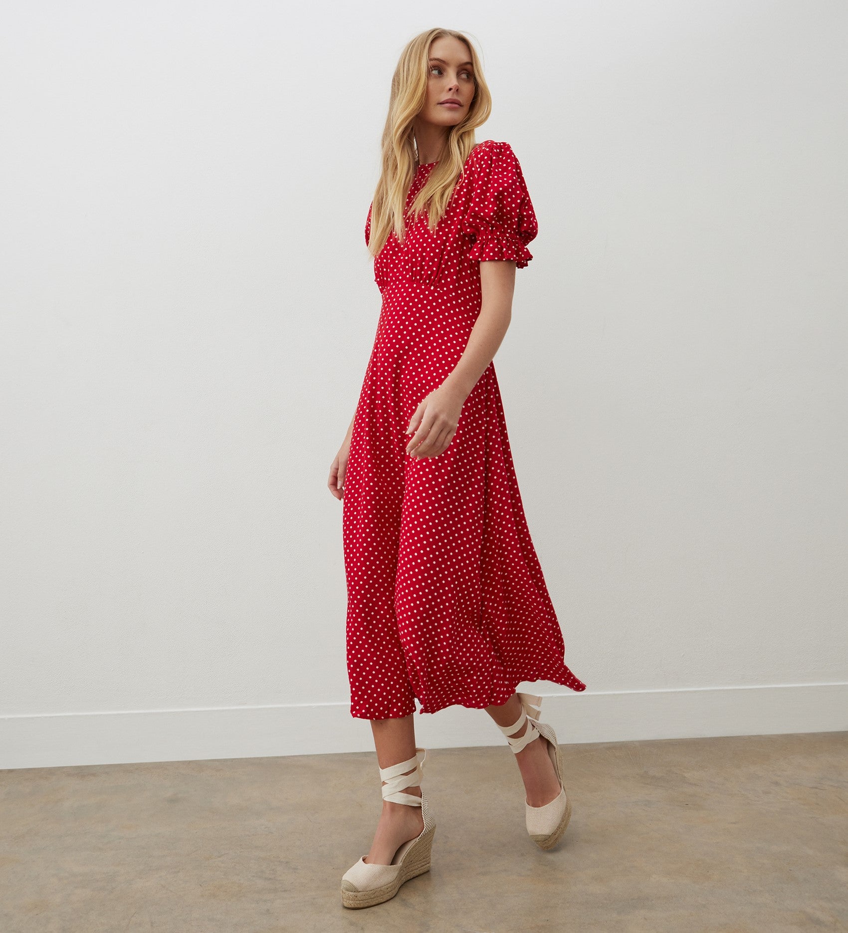 A woman in the Mela Red Polka Dot Crepe Midi Dress stands against a white wall and concrete floor, looking to the side.