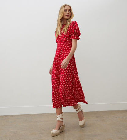 A woman in the Mela Red Polka Dot Crepe Midi Dress stands against a white wall and concrete floor, looking to the side.